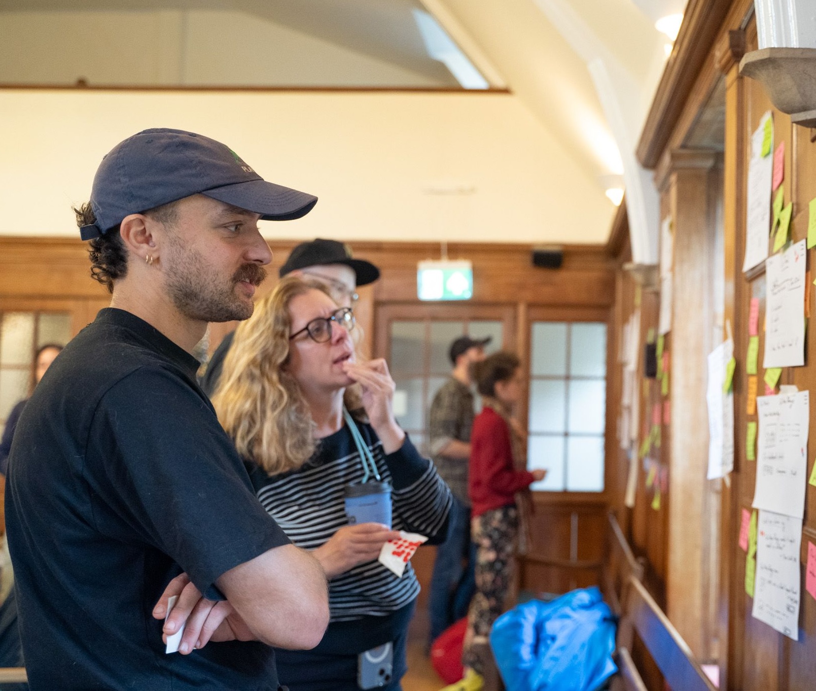 Photo from a workshop event showing several people reading sticky notes attached to a wall